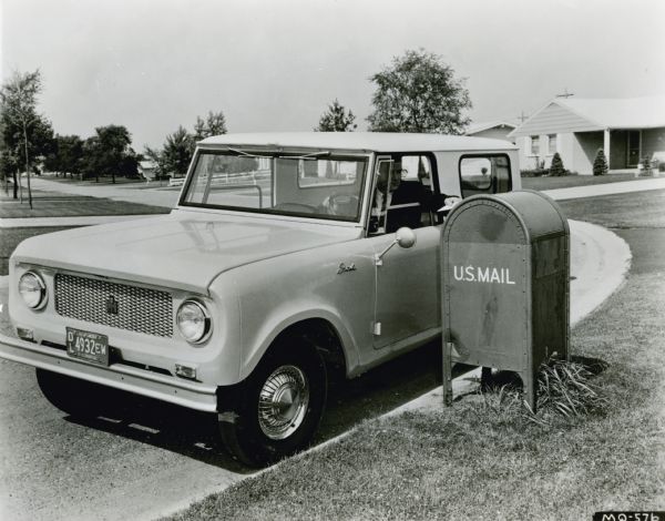 Three-quarter view from front of driver's side of an International Scout parked by mailbox. A man in the driver's seat is putting a letter into the mailbox. Original caption reads: "Photograph No. mq572. Roll-down windows for the Scout have been announced as a factory option. New windows have a wing vent and solid glass pane that retracts into the door. Advantages of the roll-down window include improved cab ventilation, cab security and driver comfort, plus a wider open window area for delivery and personal use. Standard Scout windows are removable 2-section, horizontal-sliding type. MT-2361 9/4/62" Ad code on caption reads CR-1146-D.