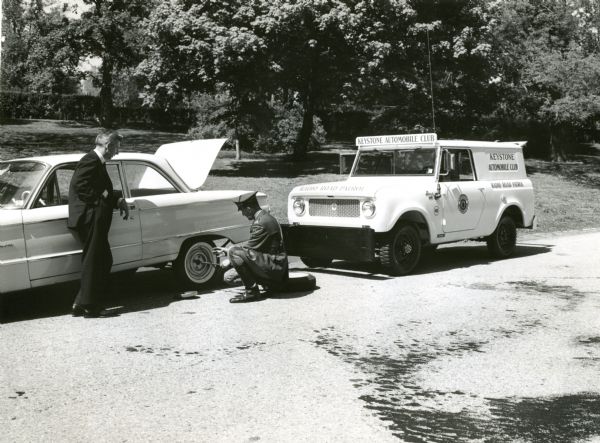 Keystone Automobile Club's International Scout used for Roadside assistance. A man is fixing the tire, while another man looks on. Original caption reads: "Photograph No. MQ-555--Patrolman of the Keystone Automobile Club's radio road patrol pulls up in his Scout emergency service vehicle to assist a member. The white Scout is one of ten purchased by the motor club to serve members primarily located in the Pennsylvania and New Jersey  area. The new additions to the organization's 25-vehicle fleet have all-wheel-drive and 93 hp. Comanche 4-cyl. engines. They are equipped with pusher bumpers, trailer hitches, and two-way radios and carry tools, auto jacks, battery re-chargers and other maintenance equipment." Ad code Cr-1146-D.