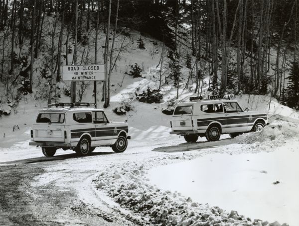 Two International Scouts entering a closed road. The roads appear to be unplowed and on a mountain. Sign reads, "Road Closed no winter maintenance." Original caption reads, "Photo TF-1344-- Versatile International Scout II utility compacts serve as official vehicles for U.S. Ski Team for 1976. The Scout's ability to negotiate unplowed mountain roads and trails while providing a smooth ride over the highway was a major feature in its selection. TD-3896."