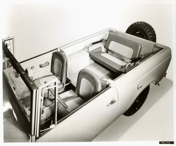 International Harvester Scout Improvement photograph. Elevated, three-quarter view from front of driver's side. Original caption reads: "Photograph MV-2518-- A major improvement in Scout body design is the removable panel in the body partition which allows easy access to the rear seat. Optional bucket seats have been redesigned for greater comfort and durability, and have two-tone heavy-duty vinyl upholstery. The two-passenger back seat, also with the same vinyl upholstery, is now mounted further back for additional leg room. Optional spare tire location outside the tailgate is also shown here. Besides these changes, the Scout also offers more efficient self-energizing duo-servo brakes, positive crankcase ventilation, greatly improved heater-defroster, and muffler mounting at rear of chassis under the body. MT-2480."