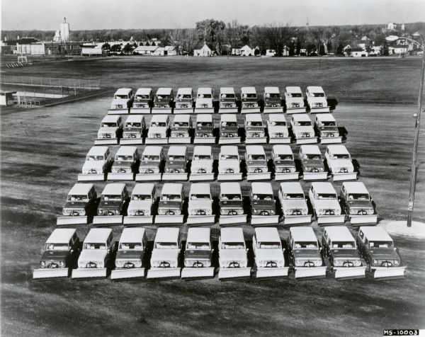 Elevated view of five rows of International Harvester Scout Snow Plows parked in a field. Original caption reads: "Photograph MS-10003-- There's muscle galore to fight winter weather in these 50 International Scouts marshalled at the International country's snow belt areas. The rugged 100-in. wb. all-wheel drive compacts feature snow plow blades with lift attachments and full-length Travel Tops. Scouts have become the first-line of defense against winter's ravages in many northern and western cities and towns. The Scout is powered by a husky 93 hp. 4-cyl. engine and is available in conventional 2-wheel drive as well as all-wheel drive. MT-2879."