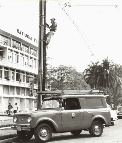 International Harvester Scout 80 used by a telephone company in Manila, Philippines. A man is climbing a telephone pole in front of a building. There is a Scout truck parked in front.