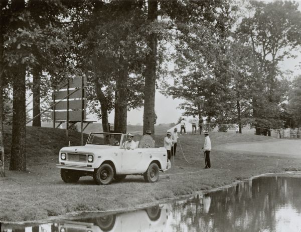 International Scout used in the American Golf Classic in Akron, Ohio. Original caption reads, "Photo #W-6632-- An unusual use for International Harvester Company's rugged Scout vehicle is its participation in the annual American Golf Classic charity event at Firestone Country Club, Akron, Ohio. Classic volunteer teams drive the loaned 800A All-Wheel Drive Scouts to handle such duties as roping and staking the course to designate spectator areas. MT-3485." Man in drivers seat of Scout, while other men string up spectator standing areas. Scout parked along the edge of a water way and trees.