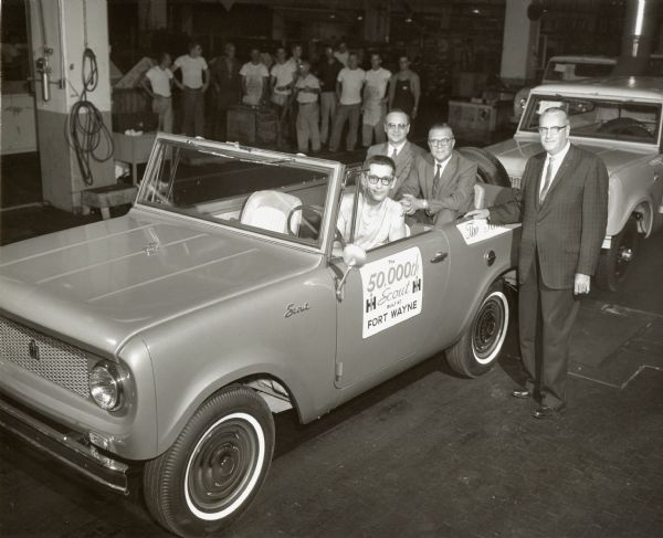 The 50,000 International Harvester Scout is rolled off the assembly line at the Fort Wayne, Indiana Works Plant. Left-frontal view. Original caption reads: "A production milestone is marked at International Harvester Company's Fort Wayne, Ind., works where the 50,000th Scout was recently produced. Checking the unit, a rear-wheel drive model with 4-passenger capacity and bucket front seats, is H.A.  Torgersen (right, works manager. In rear seat are V.A. Hart (left), superintendent of Scout assembly, and M.E. Hayhurst, works auditor, as Wayne Van Ryn, final assembly employe, drives it off the line. Introduced in January, 1961, the Scout has become the largest selling model in the International truck line. MT-2378." Sign on Scout reads: " the 50,000th Scout built at Fort Wayne." Workers seen in background of image. Factory setting.