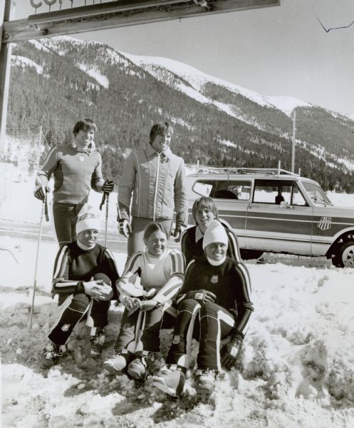 International Scout with the U.S. Ski Team. Six people pose in snow gear sitting and standing in the snow with an International Scout and mountains in the background. Original caption reads, "Photo #TF-1365-- The U.S. Ski Team recently recorded its best ever weekend of World Cup racing results at Copper Mountain, Co., as Phil Mahre (standing at left) finished second in the Giant Slalom to teammate Greg Jones. Twin brother Steve Mahre (right) took sixth in same event, second in the Slalom, and finished first in the Men's Combined Results. Susie Patterson, Lindy Cochran, (6th-Women's Giant Slalom) Billy Taylor and Gail Blackburn, (8th-Women's Slalom) (posing left to right) also participated. The team arrived at Copper Mountain in their special International Scout Four-Wheel-drive vehicles. TD-3931.