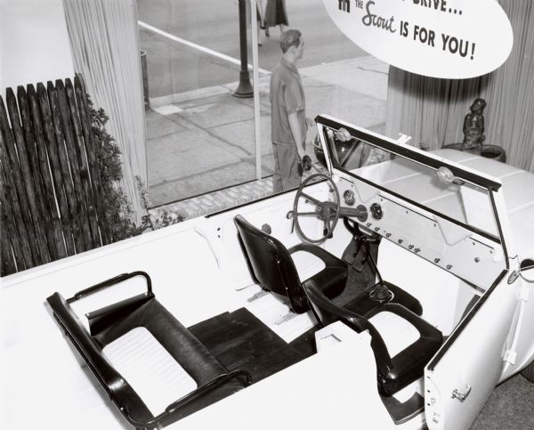 Elevated interior view of an International Scout 46 in a dealer setting. A mannequin of a man is positioned near the plate glass windows that face the sidewalk.