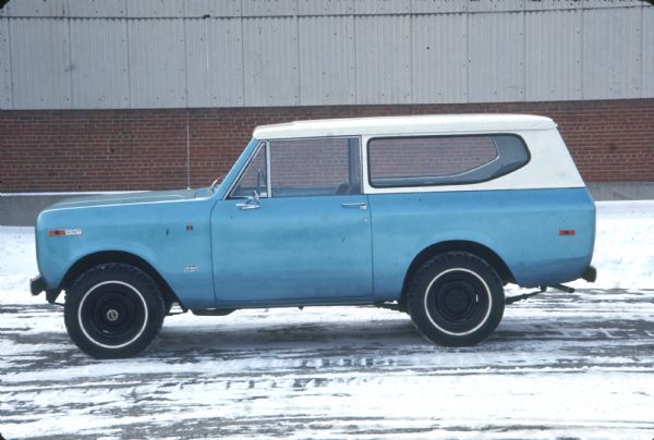 Side view of driver's side of a blue Scout with white top parked outdoors in a snowy parking area with a brick building in the background.