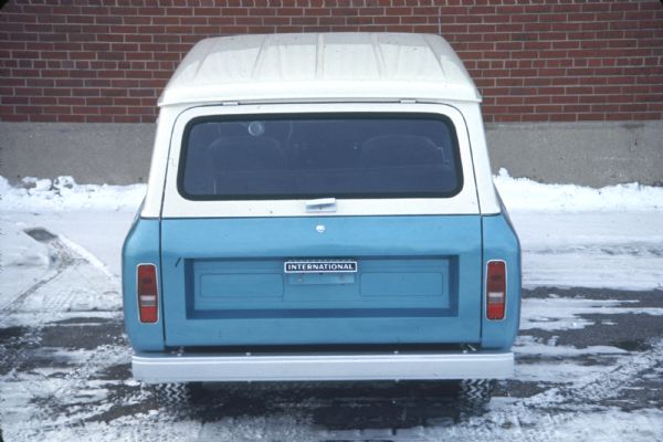 Slightly elevated rear view of blue Scout with white top parked outdoors in a snowy parking area near a brick building.