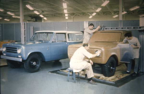 Three men indoors in a factory setting are working on a scale model of a Scout. Next to them on the left is a blue Scout with a white top.