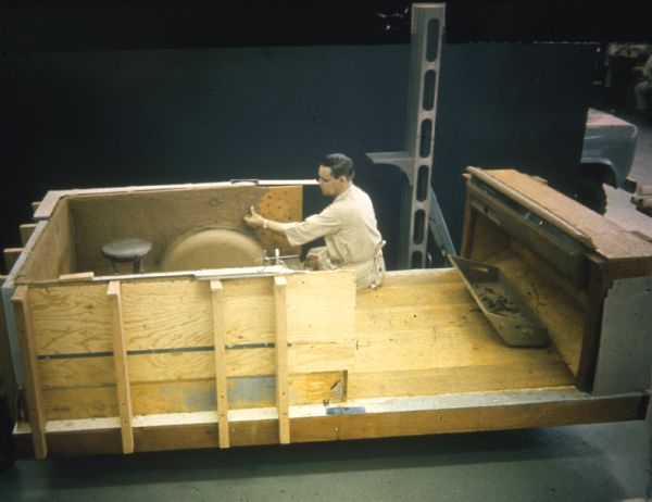 Man working on a plywood model indoors. Behind him is a wall, and on the right just behind the wall is the front hood and wheel of a Scout.