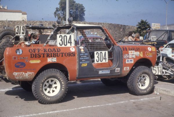 View of passenger side of orange Scout with the number "304" on the netting of the door, and behind the roll-bar. Just behind the passenger door is painted: "Driver Jimmy Jones, Co-Driver Frank Howarth, Mech. Mike McCarthy."