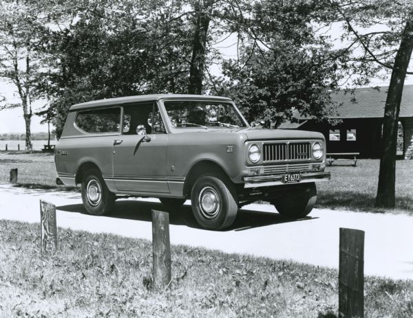 Three-quarter view from front of the passenger side of the 1973 International Scout II parked outdoors. Original caption reads: "Photo #W-6969.  With its new optional exterior trim package, the 1973 Scout vehicle is ready for both town and country. Included in the package are grille and windshield trim moulding, tail and parking lamp bezels, window frames, dual chrome outside rear view mirrors, stainless steel full wheel covers. MT-3596. 8/21/72."