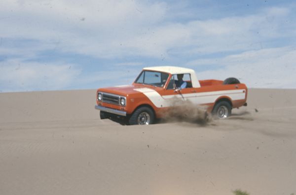 Driver's side view of Scout being driven in the sand. Orange with white detailing on side and hood. White cab top. Spare tire is attached inside truck bed.