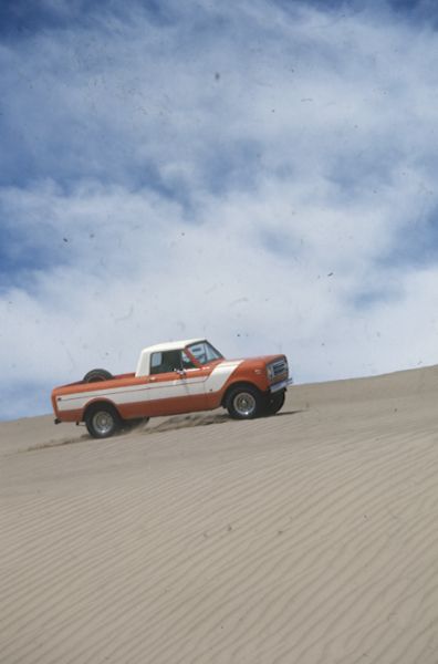 Passenger side view of Terra being driven in the sand. Orange with white detailing on side and hood. White cab top. Spare tire is attached inside truck bed.