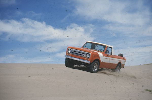 Three-quarter view from front of driver's side of Terra. A man is driving the Scout in the sand. Orange with white detailing on side and hood. White cab top. Spare tire is attached inside truck bed.