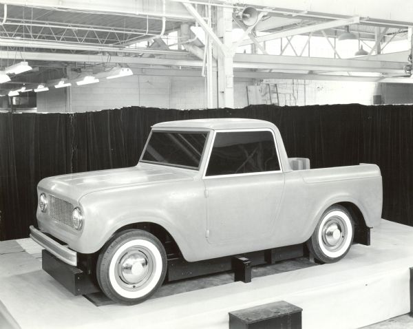 International Scout in a show room on platform. On the side panel in front of the driver's side door is a metal nameplate that reads: "Scouter." Three-quarter view from front of driver's side. Black half-curtain in background, with ceiling beams and fluorescent lighting above the curtain.