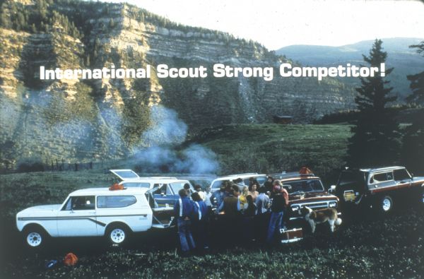 Group of people and a large dog gathered near five International Scouts parked in a grassy field. They appear to be having a cook out as there is smoke, and a cooler is sitting on the ground. There is a mountain in background.
