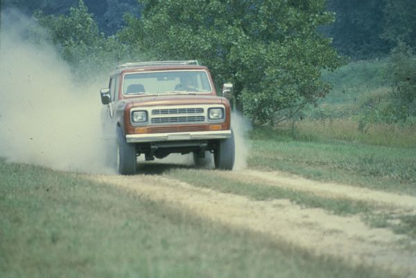 Man driving red Scout up a dusty two track dirt road through a grassy area. Hills and trees in the background.