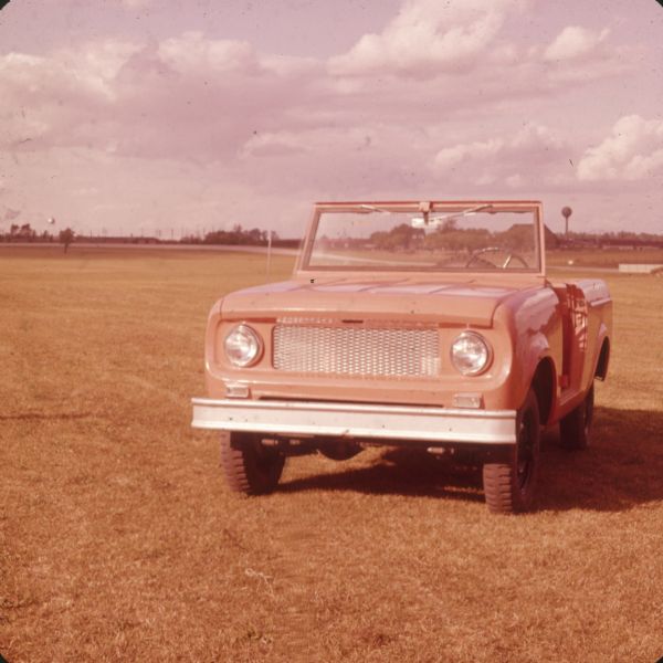 Red or orange Scout parked in a large field of mowed grass. There is a water tower in the far background.