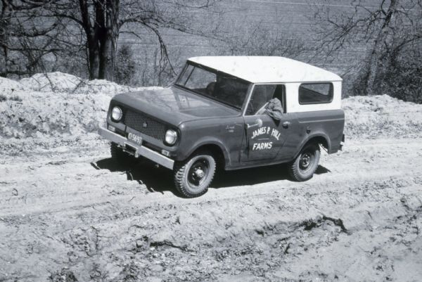 Slightly elevated view of man driving a Scout on a steep dirt road. Painted on the driver's side door is: "James P. Hill Farms." Trees line the road, and a field is below in the far background.
