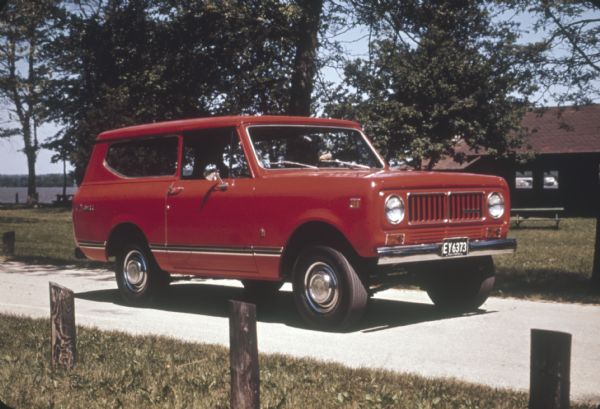 A person is driving a red Scout on a road in a park or recreation area. There is a park building in the background on the right, and a picnic table is near a lake on the left.