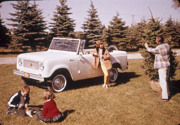 A man, woman, and two children posing with white Scout. The spare tire is mounted on the back. There is no top on the Scout. The man is standing and holding an evergreen tree, and the two children are sitting on the ground with a cat. The woman is leaning against the driver's side door of the Scout.