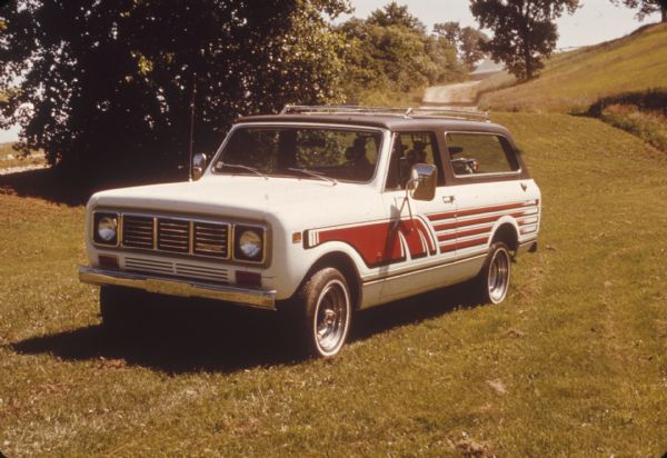 Three-quarter view from front of driver's side of a white Scout with red detailing parked in the grass. Trees and dirt road in the background.