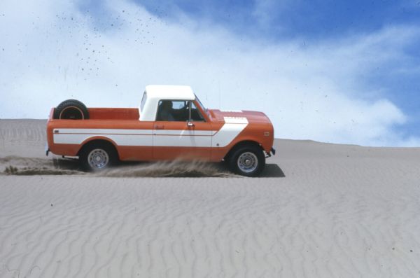 Passenger side view of Terra being driven in the sand down a steep hill. Orange with white detailing on side and hood. White cab top. Spare tire is attached inside truck bed.