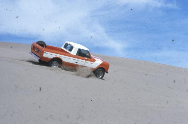 Passenger side view of Terra being driven in the sand down a steep hill. Orange with white detailing on side and hood. White cab top. Spare tire is attached inside truck bed.
