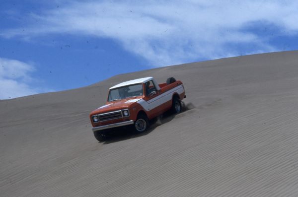 Three-quarter view from front of driver's side of Terra. A man is driving the Scout down a steep hill in the sand. Orange with white detailing on side and hood. White cab top.