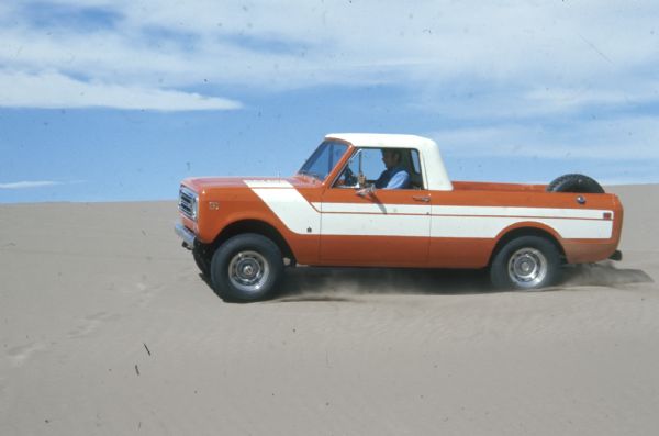Side view of driver's side of Terra being driven in the sand. Orange with white detailing on side and hood. White cab top. Spare tire is attached inside truck bed.