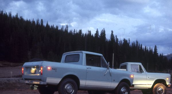 Blue Scout II XLC parked outdoors. Another truck is behind on the right. Forested hill in the background. Snow-capped mountains in the far background.