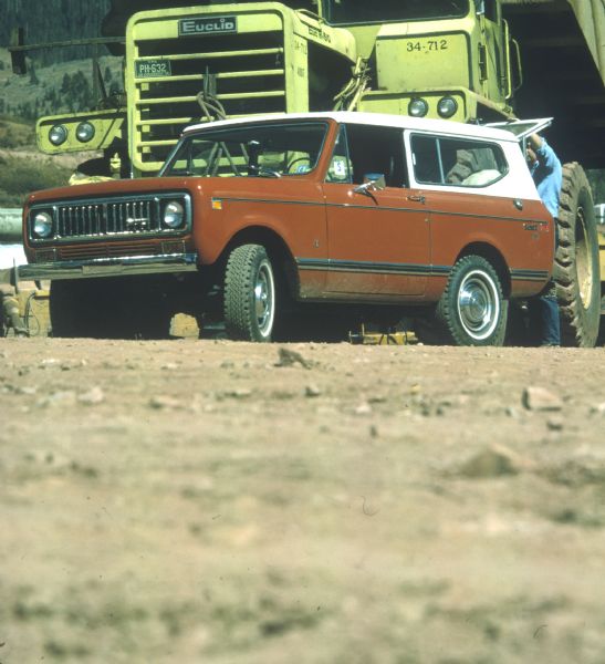 Red/orange Scout II XLC parked in front of a large construction vehicle. A man stands holding open the hatchback. Mountain in the background. 