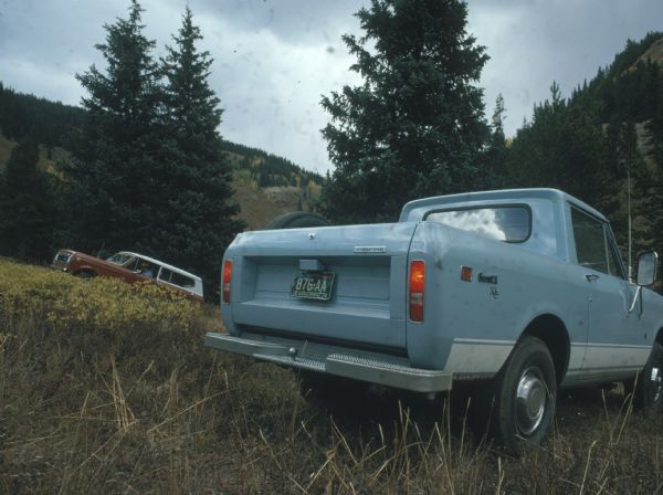 Three-quarter view from rear of blue Scout II XLC parked on steep hill in a grassy field. In the background on the left a man sits in another Scout, red/orange with white cab top.