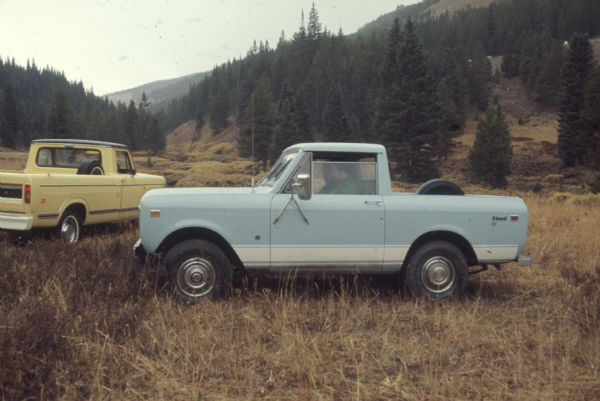 Two men sit in a blue Scout II XLC in a field in a mountainous area. A yellow truck is parked behind them on the right.