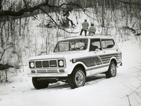 International Scout in the foreground. In the background are two hunters walking into the woods in the snow on a hill. 