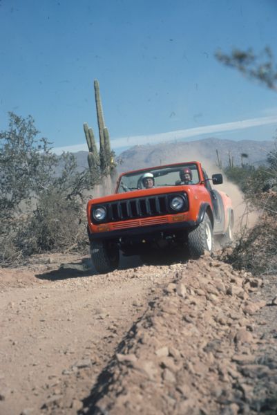 Three-quarter view from front of driver's side of Red Scout. The Scout is being driven up a dirt road. Cactus and mountains in the background. Both driver and passenger are wearing helmets. 