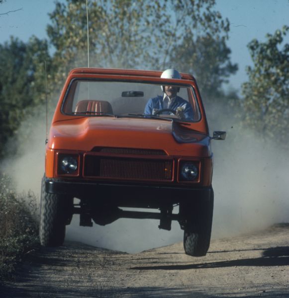 View looking down dirt road of a man wearing a helmet driving a red Scout. All four wheels are in the air.