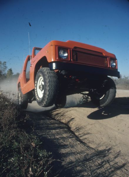 Three-quarter view from front passenger side of a red Scout being driven up a steep dirt road. Most of the wheels are in the air.