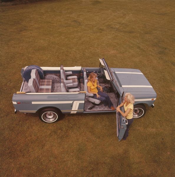 Overhead view of two women with a Scout parked in a grassy field. Blue/grey with white stripe detailing. Grey seats with plaid. One woman sits in the swivel passenger seat, the other woman leans on the open passenger door. Top has been removed.