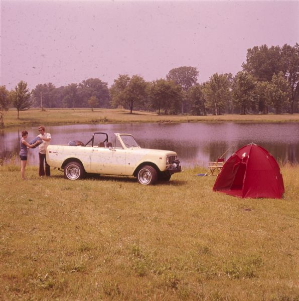 A man and a woman are standing in the grass near a small body of water and a white colored Scout II XLC. Near the parked vehicle is a red tent and a chair with a fishing pole resting on it. Trees are in the background behind the lake or pond.