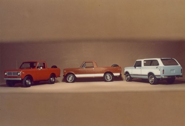 Driver's side view of three Scouts parked indoors in a half circle. On the left is a red Scout with a spare tire mounted in the open truck bed. In the center is a brown Scout with white detailing, and a spare tire mounted in the open truck bed. On the right is a blue Scout, with white detailing and white cab rood and truck bed topper.