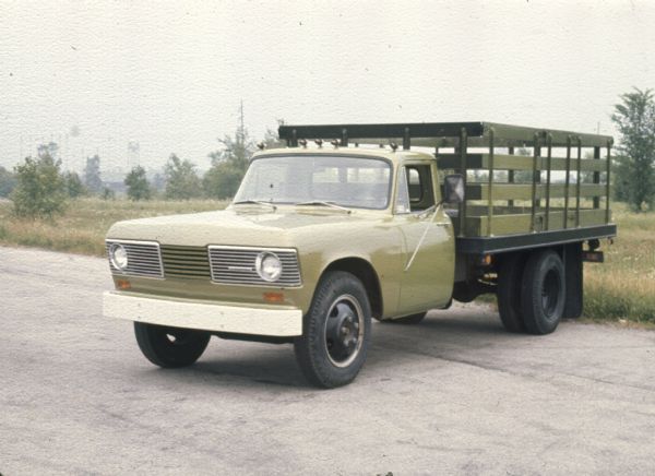 Three-quarter view from front of driver's side of tan truck with stake body parked outdoors.