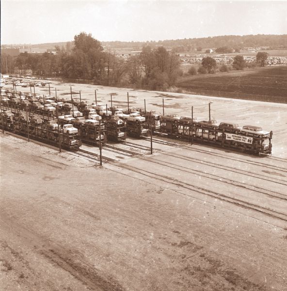 Elevated view of "Texas Special" train load of light duty vehicles at a railhead. There are railroad cars on five tracks loaded, double-decker style, with vehicless. Banner on the front of the far right set of railroad cars has a banner that reads: "IH Trucks for Houston, Texas."