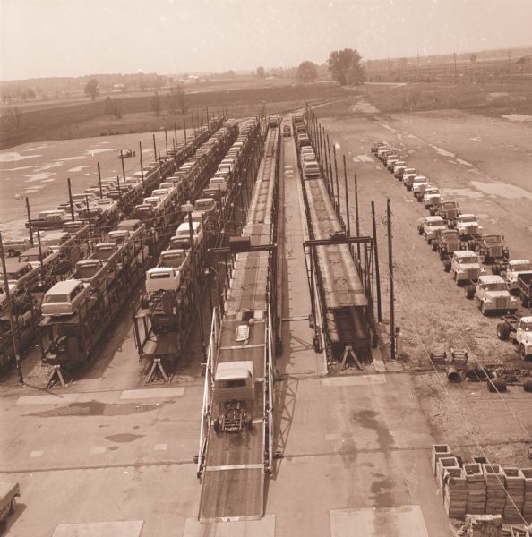 Elevated view from the rear of "Texas Special" train load of light duty vehicles at a railhead. There are railroad cars on five tracks loaded, double-decker style, with IH trucks. The trucks appear to be in the process of being loaded, with one truck on the ramp up to the second level. A row of trucks are parked to the right of the tracks, ready to be loaded.