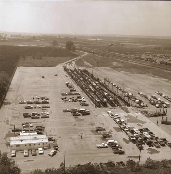 Elevated view of railhead. Cars and trucks are parked near small industrial buildings on the bottom left. The "Texas Special" train load of light duty vehicles are on five railroad tracks, and are loaded, double-decker style.