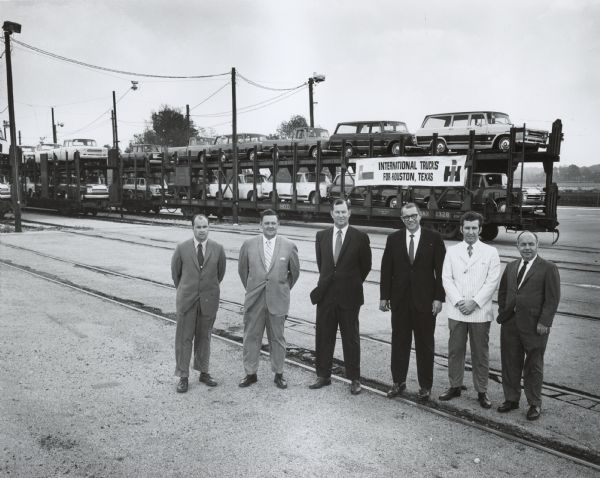 Group of six men wearing suits posing in front of "Texas Special" train load of light duty vehicles at a railhead. There are railroad cars on five tracks loaded, double-decker style, with vehicles. Banner on the front of the far right set of railroad cars has a banner that reads: "International Trucks for Houston, Texas."