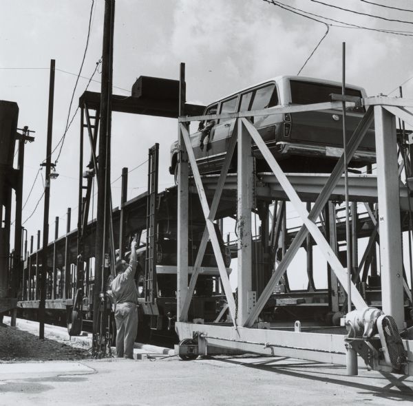Man working at the side of a railroad car on the "Texas Special" train load of light duty vehicles at a railhead. Another man is driving a vehicle up the ramp to the second level, as the railroad cars are loaded, double-decker style.