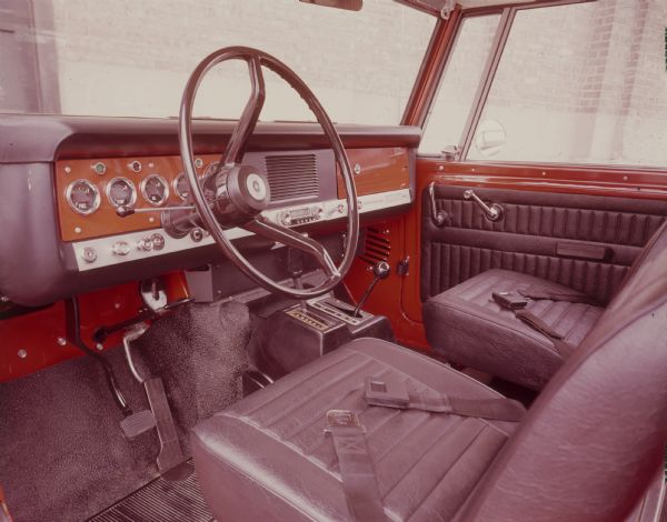 Interior view of the driver's seat, steering wheel, and dashboard of Scout. "International Scout 800" is printed on a panel under the glove compartment in front of the passenger seat.