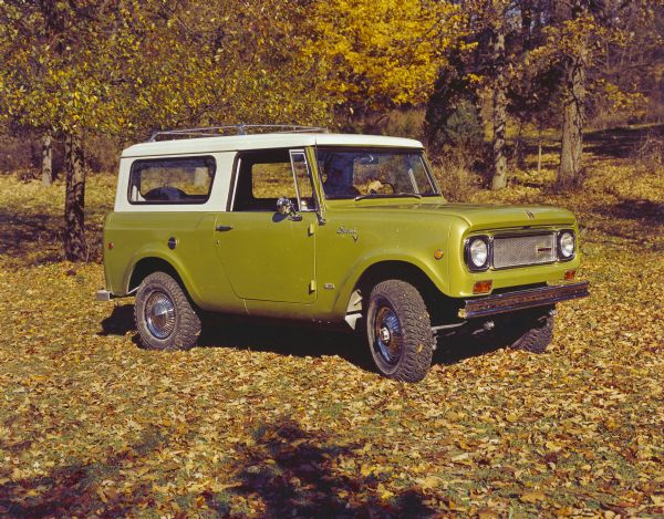 Three-quarter view from front of passenger side of Green Scout with white top. There is a rack on top of the roof and a spare tire mounted on the back. Insignia on side read: "IH," "Scout," and "ALL WHEEL DRIVE." A man is sitting in the driver's seat. Fall leaves are on the trees in the background.
